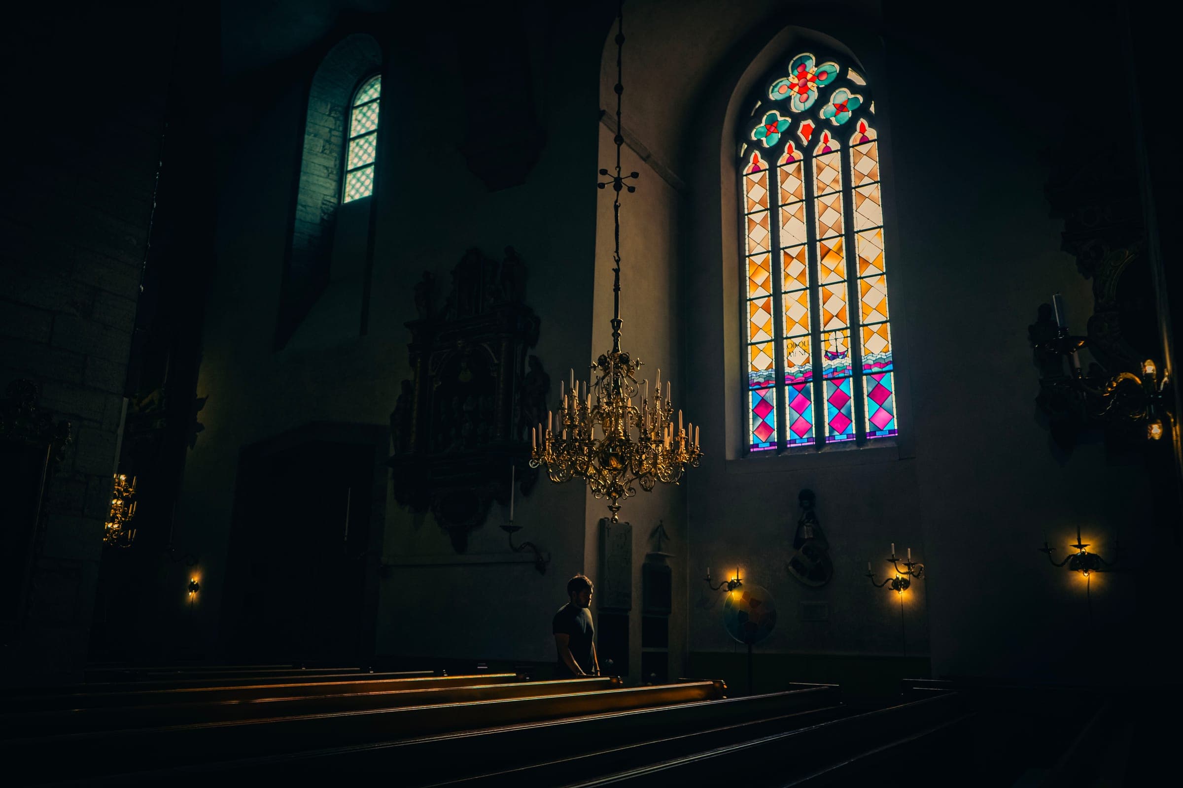Kyrkorum i dunkelt ljus med gyllene ljuskrona och färgstarkt blyinfattat fönster
