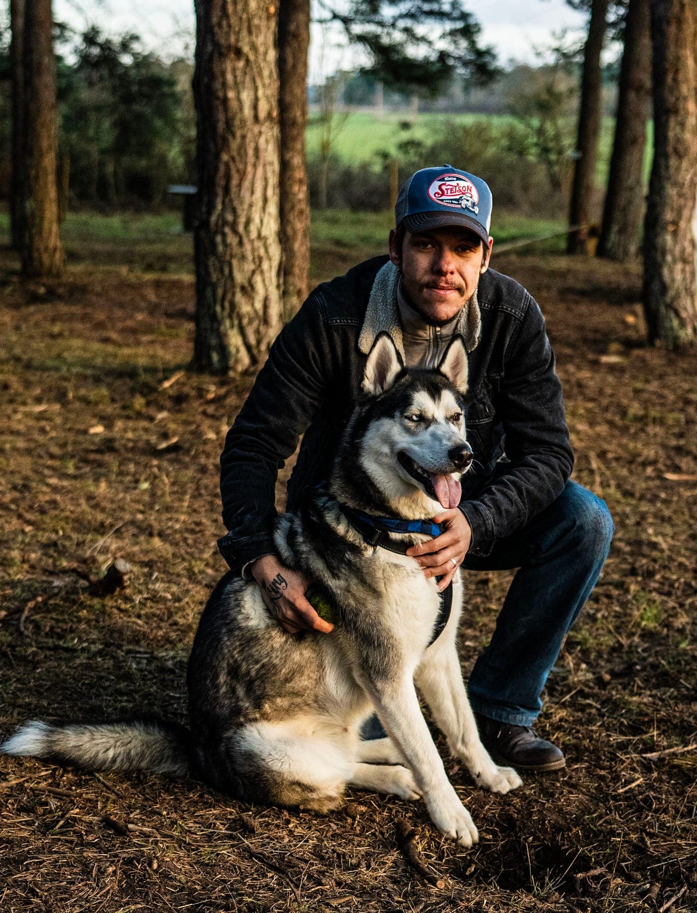 Man i keps sitter på huk bredvid en husky i skogen i skymningsljus